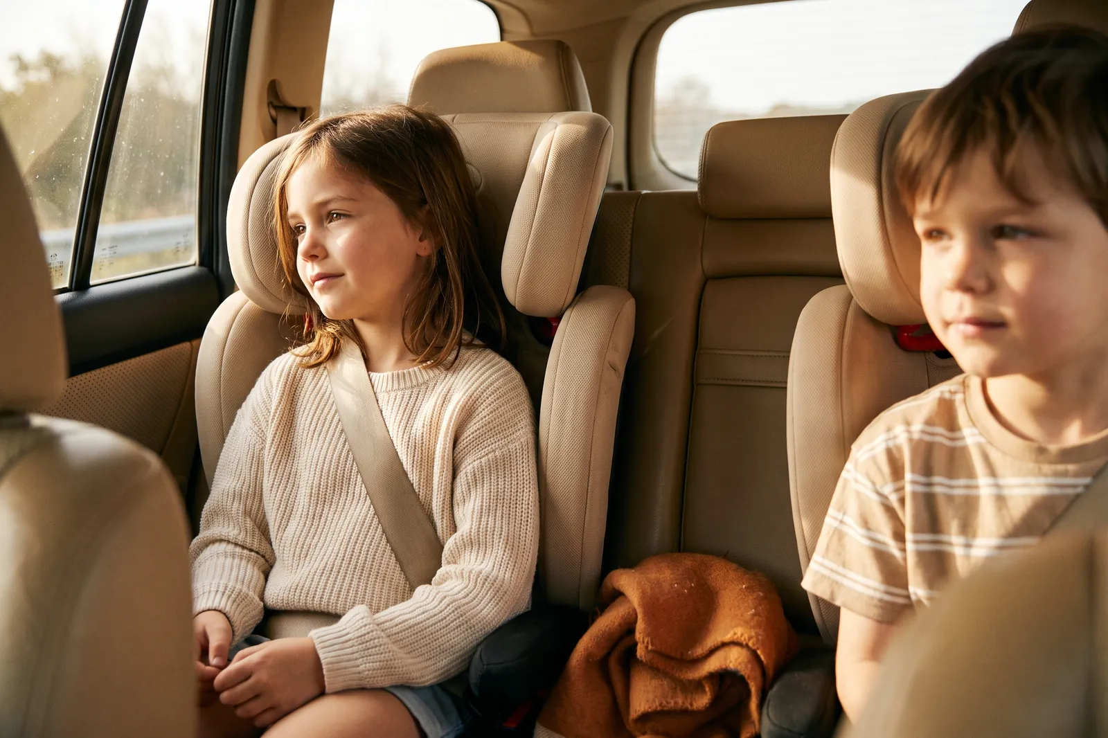 Two siblings in booster seats listen quietly in the back seat of a car on a sunny afternoon