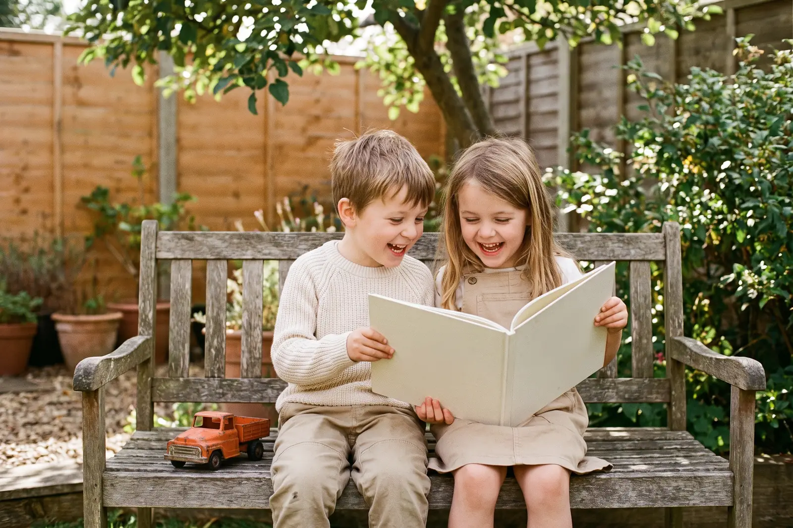 Two young friends share a picture book on a backyard bench, laughing
