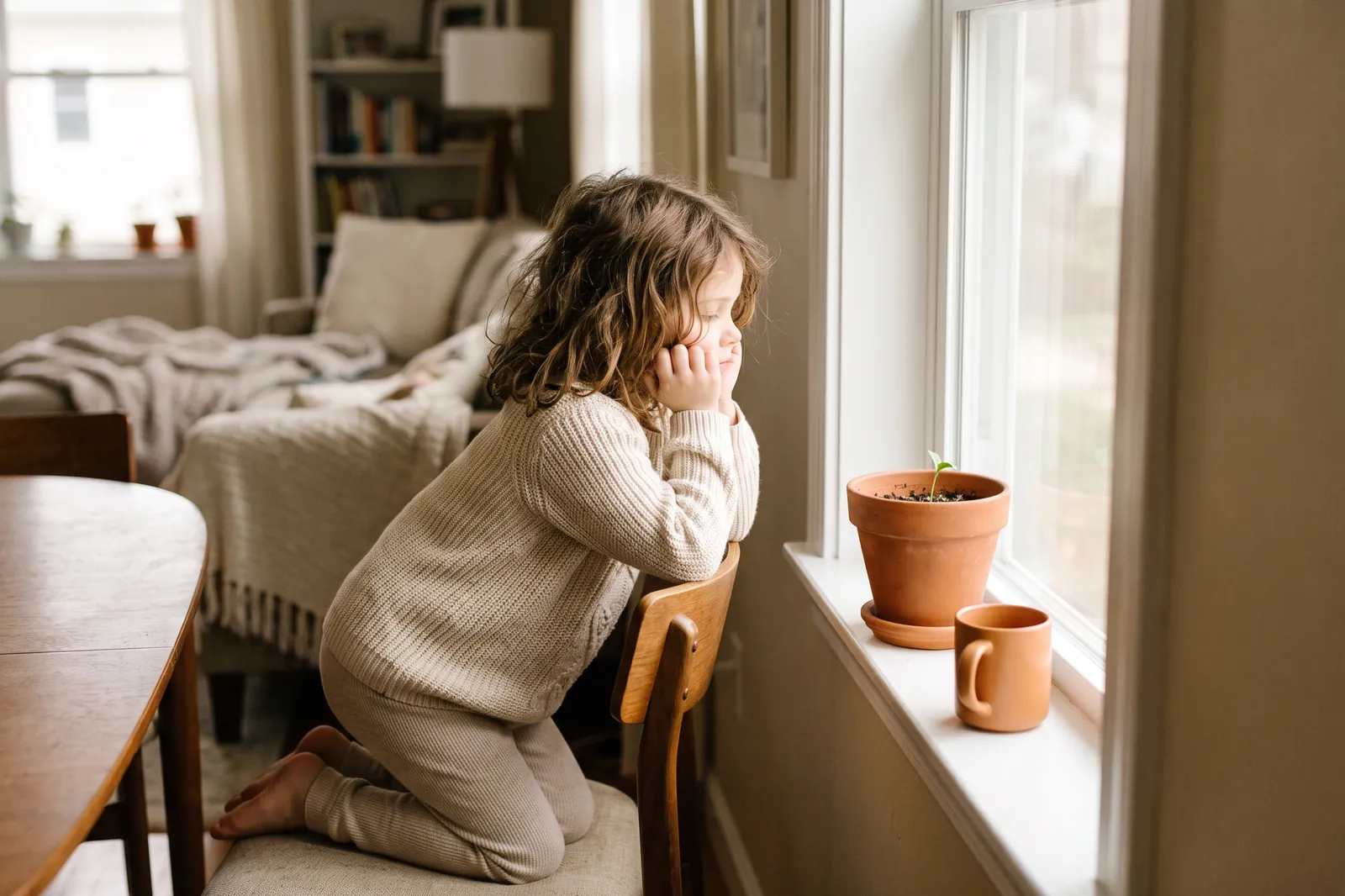 A young girl rests her chin on her hands, watching a seedling sprout in a pot on the windowsill