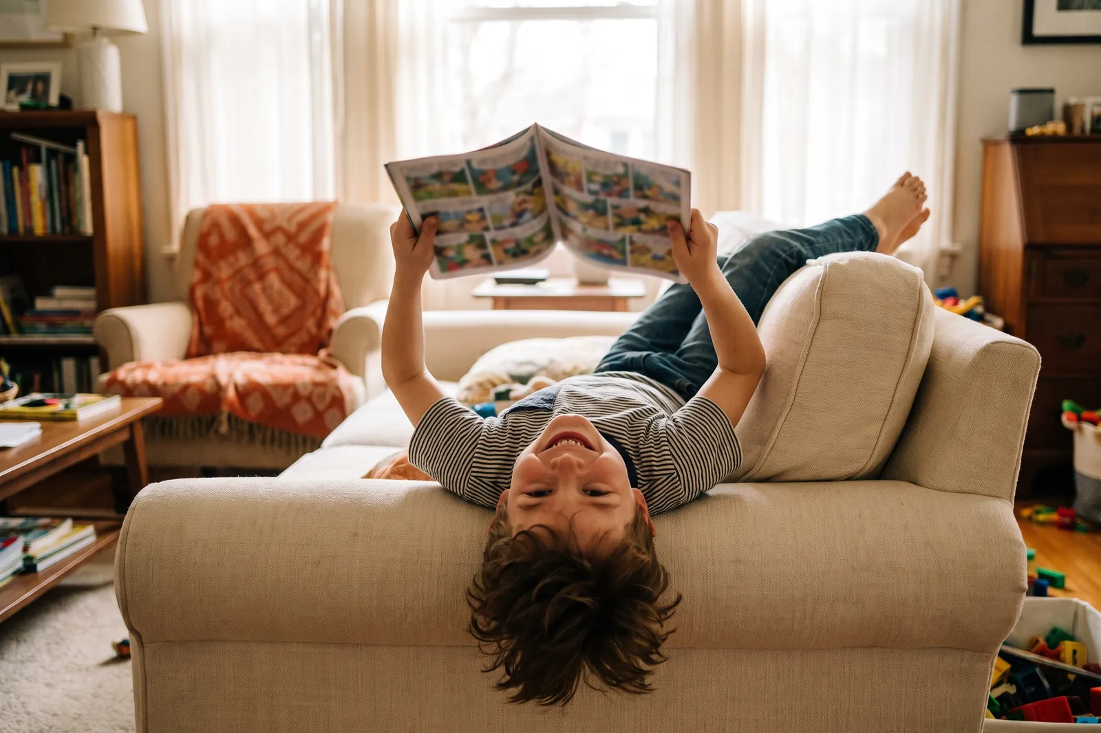 A boy lies upside down on the sofa grinning at a comic-style book