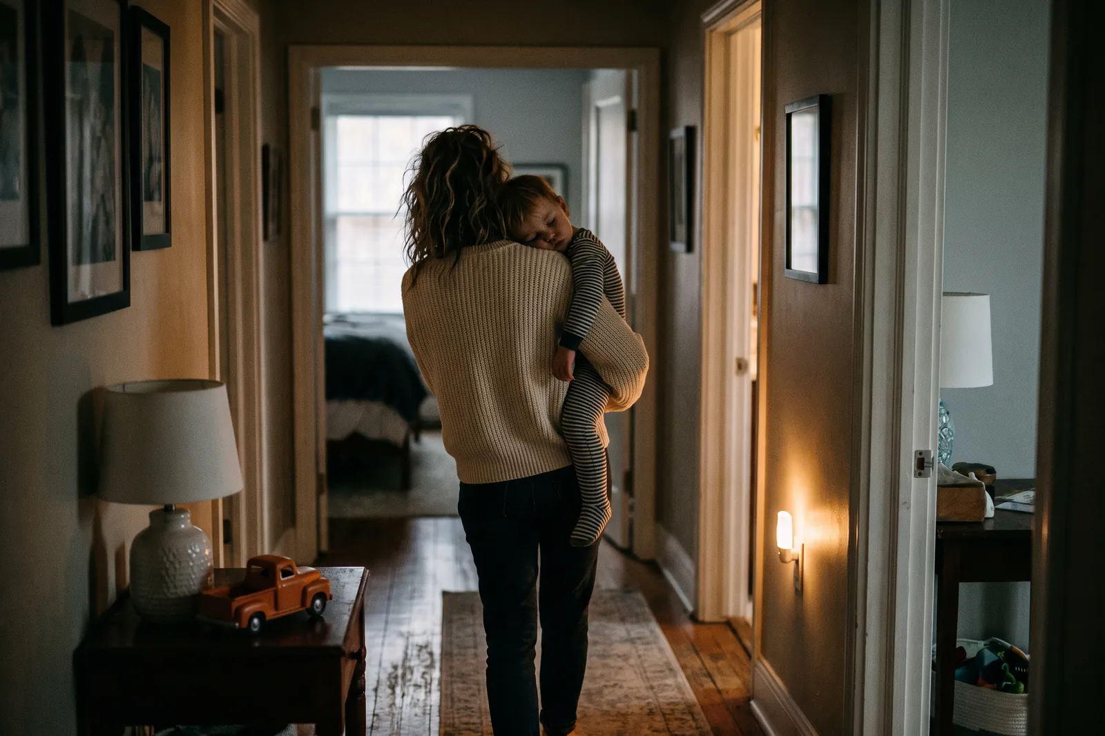 A mother carries her sleepy toddler down a dim hallway at night